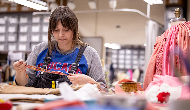 Josie Everett stands before a table scattered with fabrics. She is looking down at a piece of fabric she’s holding in her hands. Beside her is a pink skirt draped on a mannequin.