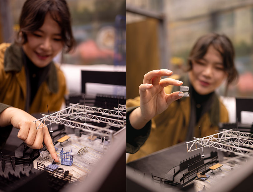 (Left) A Inn Doo points at a tiny dolly model within her ¼-inch scale model of the Barber Theater. (Right) A Inn Doo holds a miniature shopping cart between her index finger and thumb above a ¼-inch scale of the Barber Theater.
