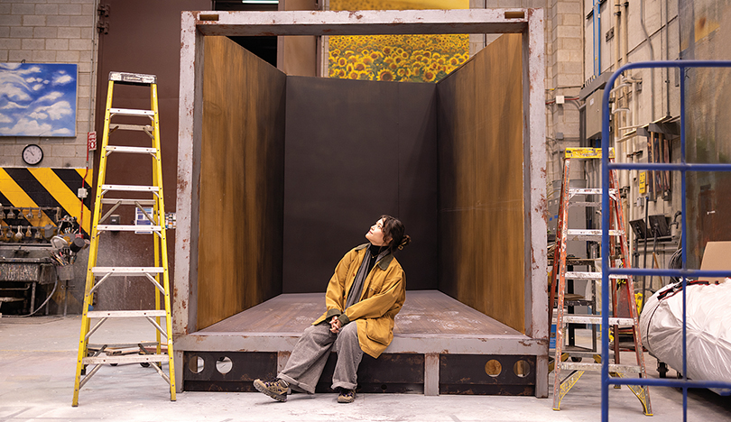 Doo sits in a large wooden box in the Wirtz Center’s scenic design shop, looking up at the ceiling.
