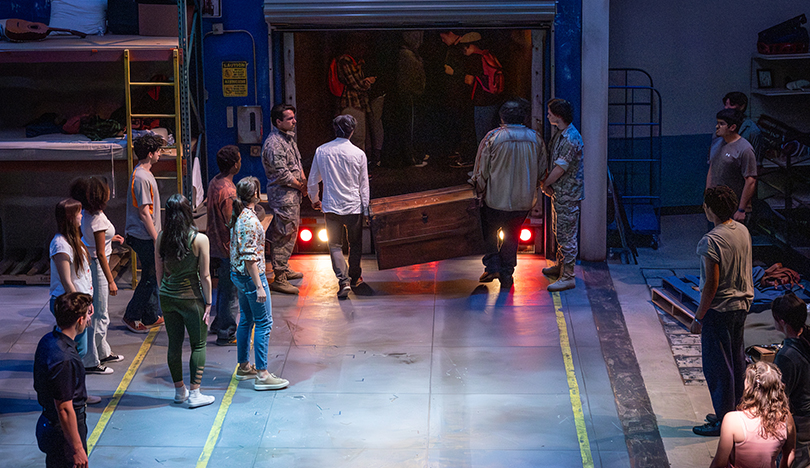 Onstage, Cervantes and his friend hold either end of a large wooden chest as they prepare to step into the back of what appears to be a truck. Other detainees look on from the sidelines.