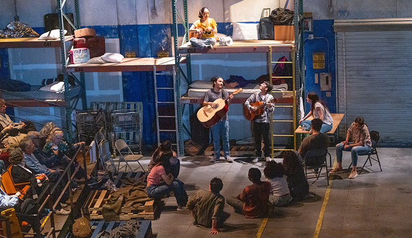 A view of the stage with distressed-looking walls, rickety shelving with ladders, a garage door, pallets, and actors on stage. The audience is visible on the lefthand side.