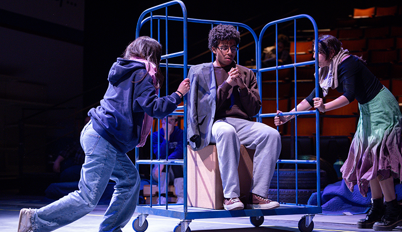 An actor sits on a box on a dolly with their hands folded in prayer while two other actors push the dolly on either side.