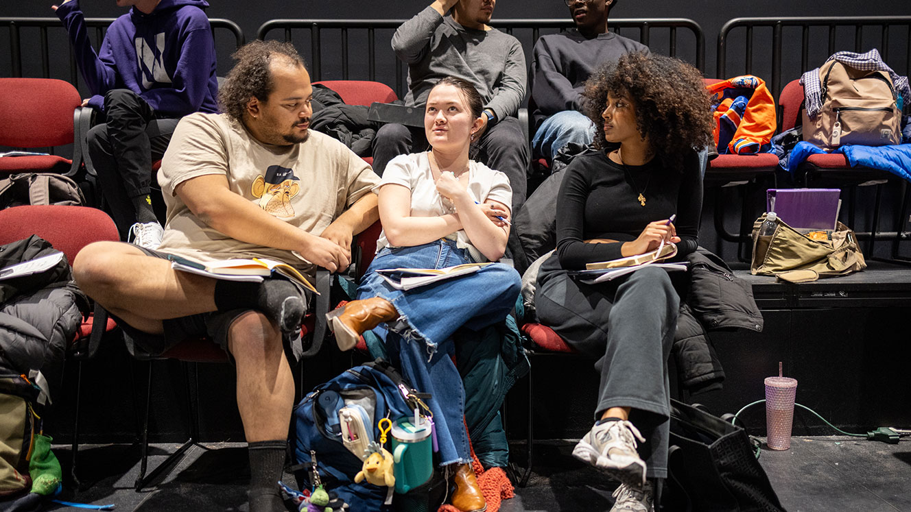 Three students sit together in the block box theater with scripts on their laps.