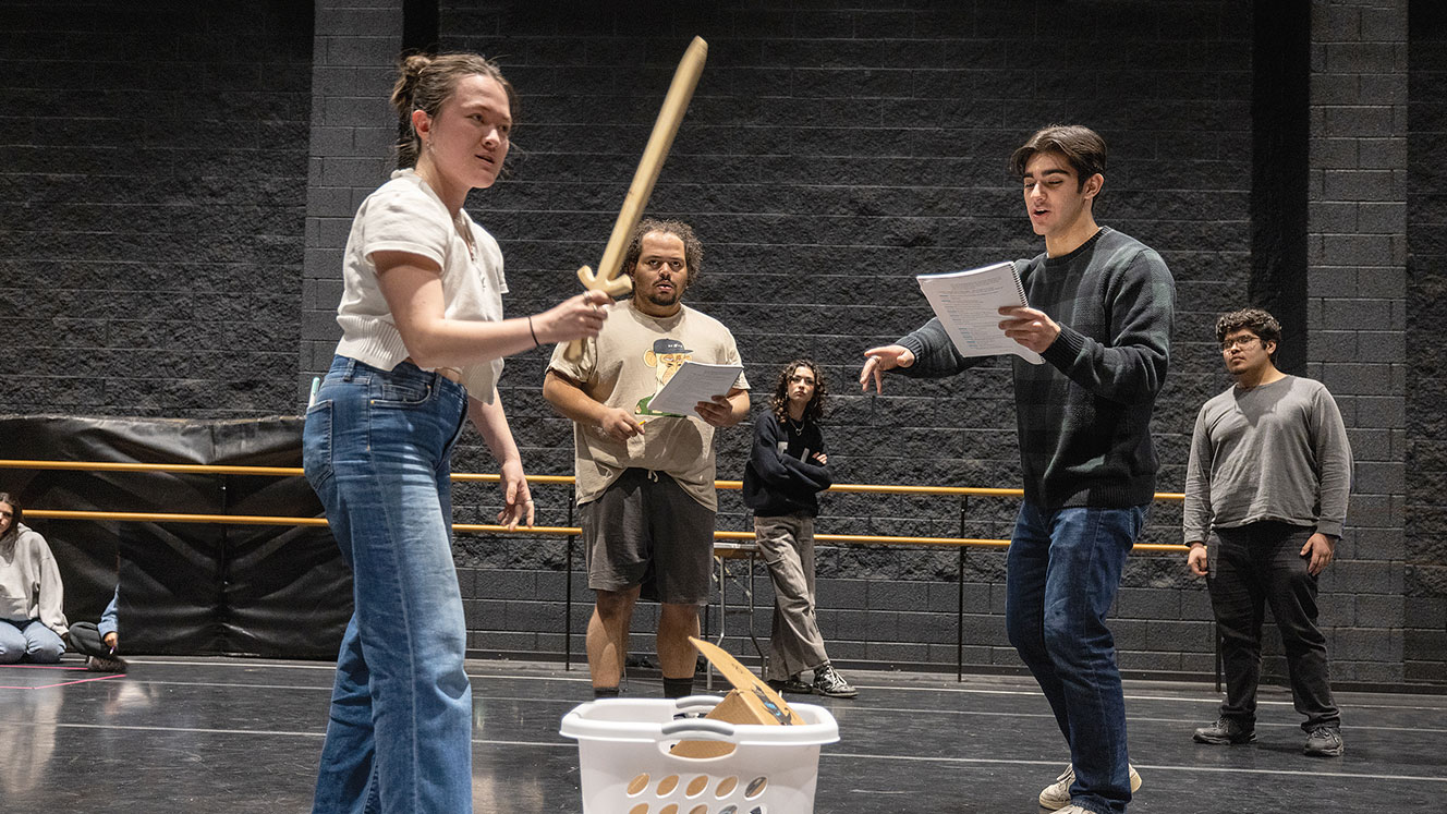 Actors stand in a black box theater blocking out a scene using props. A person in a white top and blue jeans holds a sword, body tilted toward the camera. A laundry basket is on the ground in front of them. A person in a striped sweater and blue jeans gestures toward the actor with the sword, holding a script in their other hand. Another actor stands in the back holding a script, while two additional people watch. 