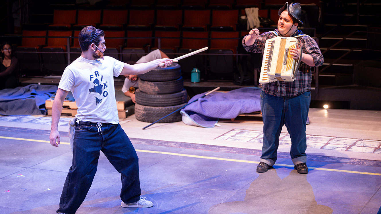 Two actors stand on stage in the Barber Theater. One points a wooden sword at the other, who wears a helmet and carries an accordion.