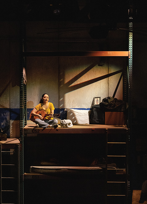 Alexa Núñez Magaña sits on a lofted wooden plank, playing the vihuela while singing.