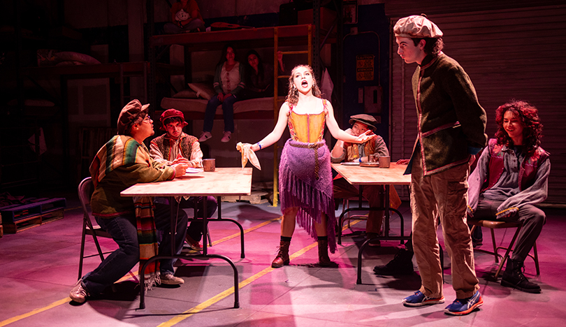 Coco Gonzalez stands on stage, gesturing toward the audience with her hands while singing. There are tables and other actors on either side of her.