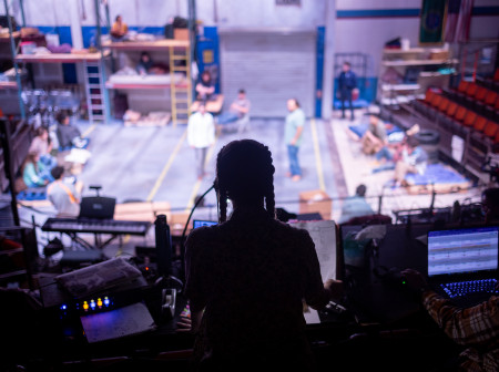 A view from up in the seating area of the Ethel Barber Theater, looking down at the stage, where actors are rehearsing. The stage is lit with white light, and the actors are out of focus. In the foreground is a backlit silhouette of Audrey Michael sitting at a table with computer screens and gadgets. 