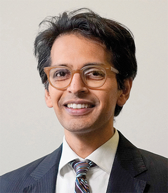 Raj Mankad smiles for the camera wearing a white shirt, neck tie and dark suit jacket and glasses