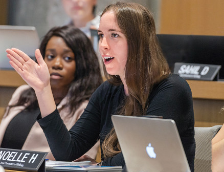 A student speaks with her hand raised at the Kellogg Climate Conference, as two students watch her in the background. 