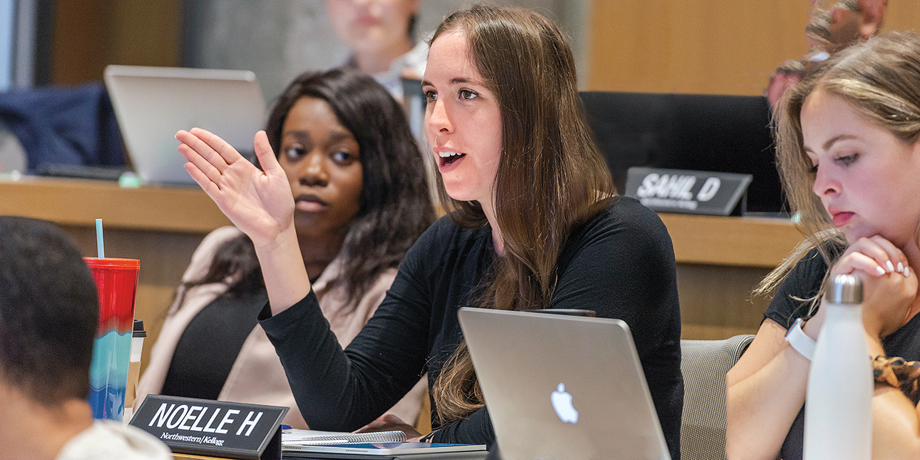 A student speaks with her hand raised at the Kellogg Climate Conference, as two students watch her in the background. 