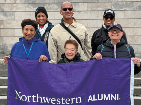Six Northwestern alums stand on the steps of a building in Paris holding a purple flag that reads, “Northwestern Alumni.”