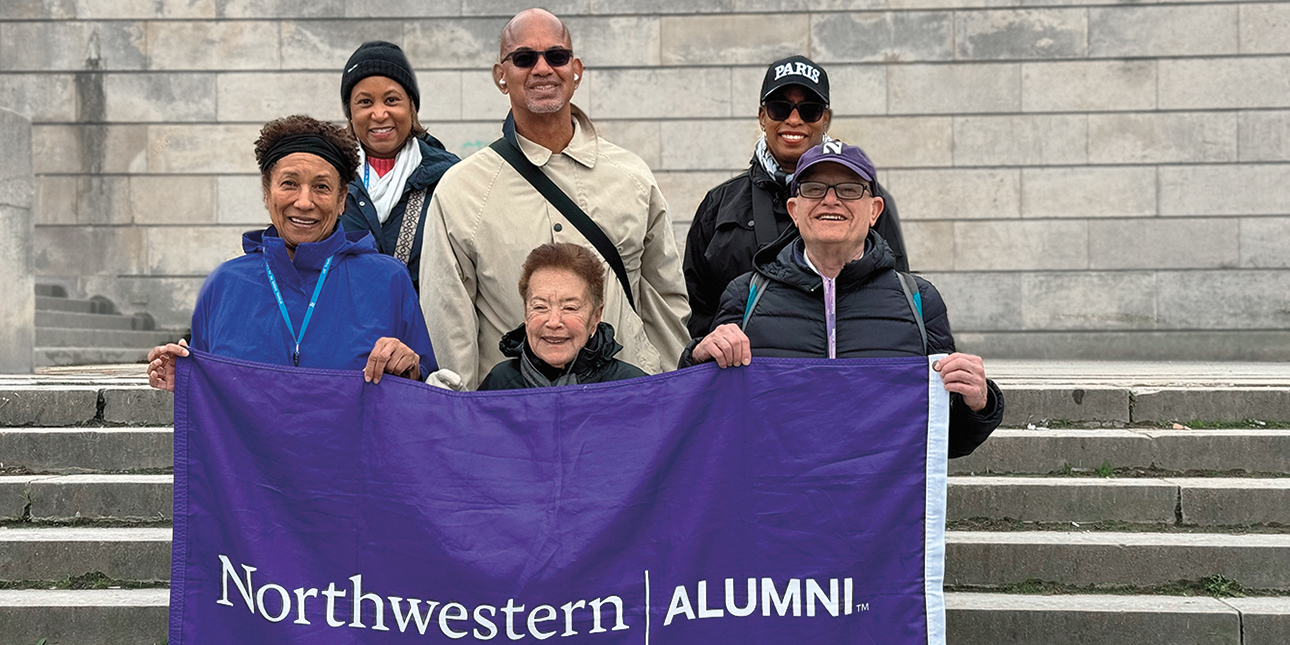 Six Northwestern alums stand on the steps of a building in Paris holding a purple flag that reads, “Northwestern Alumni.”