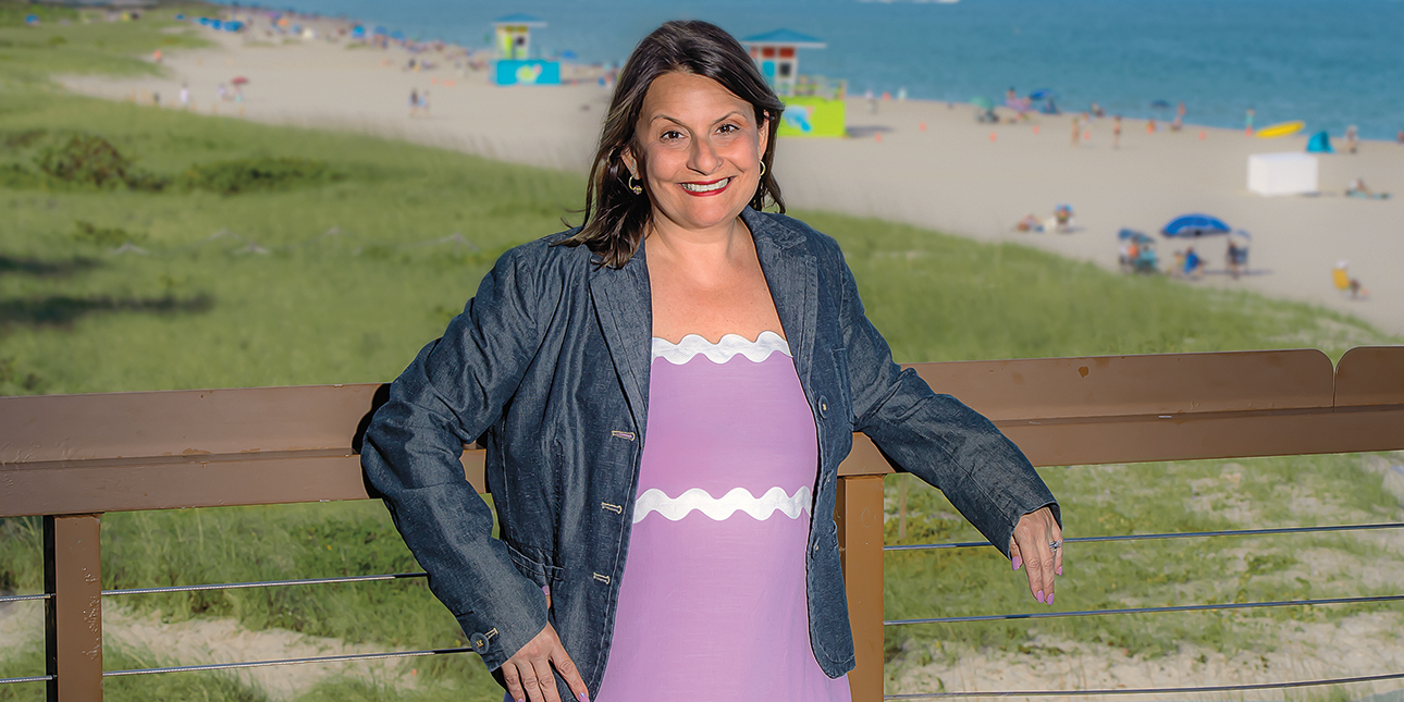 Maria Atanasov poses on a boardwalk with her arms propped up on a wooden railing in front of a sunny beach in Florida.