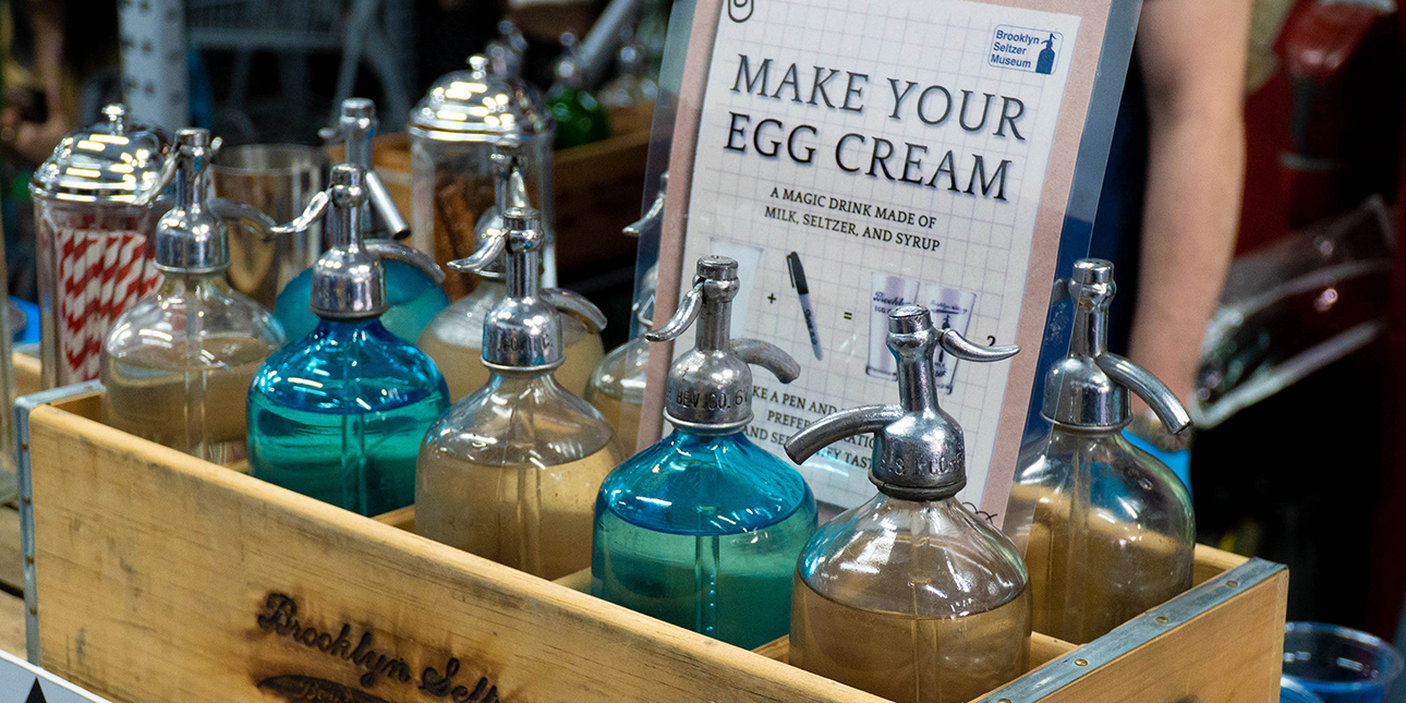 A wooden crate holds blue and tan bottles with ingredients to make an egg cream.