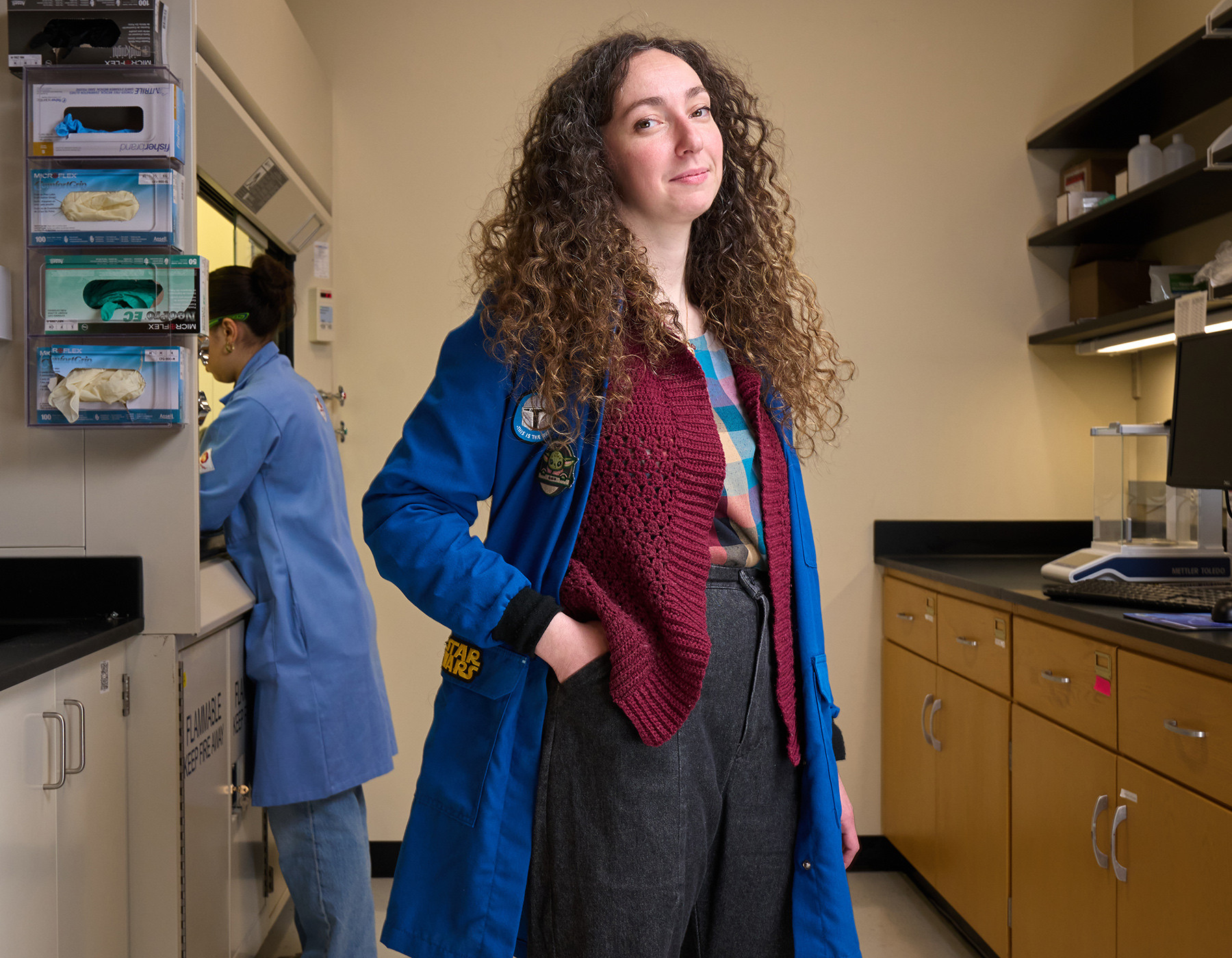 Cécile Chazot stands smiling, facing the camera, with a blue lab coat over her burgundy sweater and black jeans. 