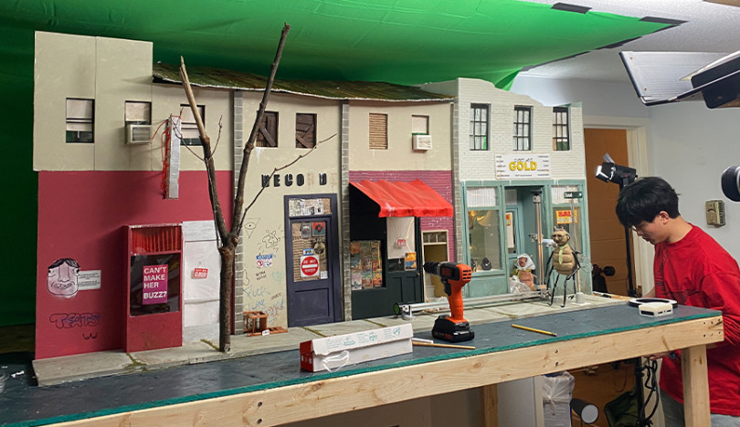 A cardboard-constructed street scene stretching roughly 8 feet from the film Common Fly is displayed on a table with a green screen behind it. A student adjusts a corner of the scene. A power drill can be seen on the table.