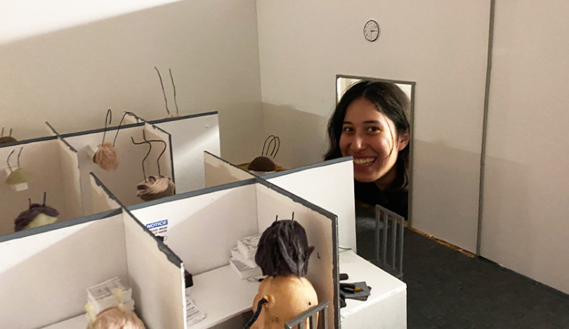 A student sticks her head through the doorway of a cardboard-constructed office scene. Bug antennae can be seen poking out from cubicles.