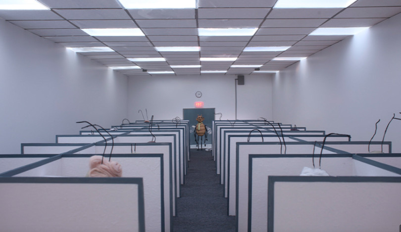 A fly model stands in the middle of a drab office where bug antennae stick out from cubicles.