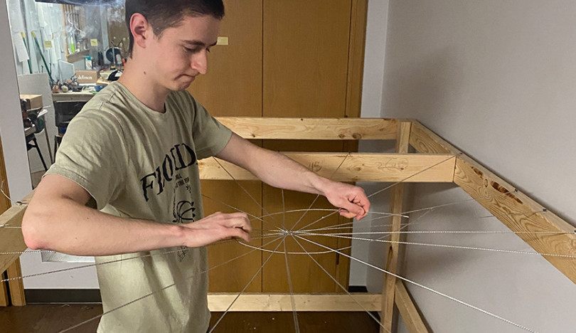 A student stands in the middle of a large web of wires held up by wooden scaffolding. He is arranging the pieces into a spiderweb formation. 