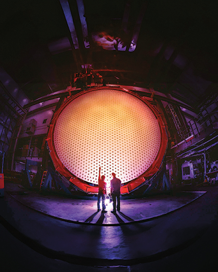 Two people, one with their arm raised up in the air, stand in front of a giant, orangey-yellow circular mirror, one of the seven primary mirrors of the GMT.
