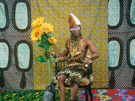 A Black man draped in gold jewelry and leopard print clothing wrapped around his waist sits in a leopard-print chair, holding sunflowers, against a colorful patchwork background.