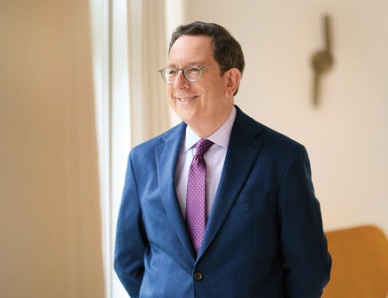 Michael Schill, wearing a blue suit, purple polka-dot tie and glasses, is smiling and looking away from the camera, with his hands behind his back and his body turned slightly to the left. 