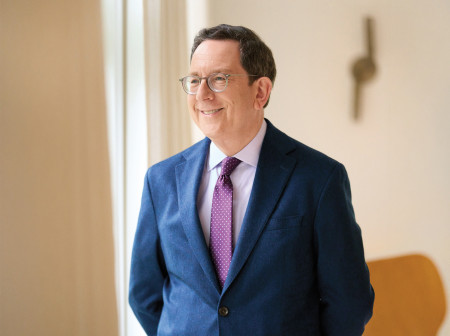 Michael Schill, wearing a blue suit, purple polka-dot tie and glasses, is smiling and looking away from the camera, with his hands behind his back and his body turned slightly to the left. 