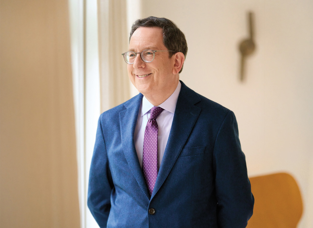 Michael Schill, wearing a blue suit, purple polka-dot tie and glasses, is smiling and looking away from the camera, with his hands behind his back and his body turned slightly to the left. 