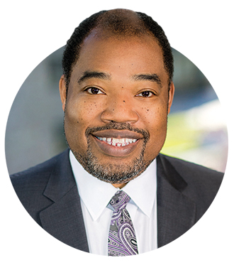 Anthony Chambers is smiling at the camera. He is wearing a dark suit, white shirt and paisley purple, white and black tie.