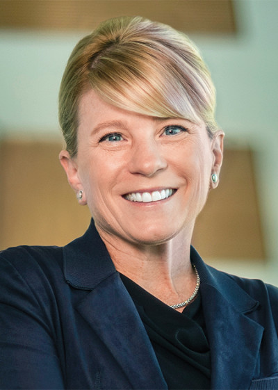Shana Kelley, wearing a black suit and her hair up in a bun, smiles at the camera in front of a brown and white wall.