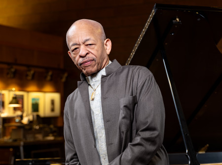 Steve Colson wears a gray suit and leans back against a Steinway grand piano.
