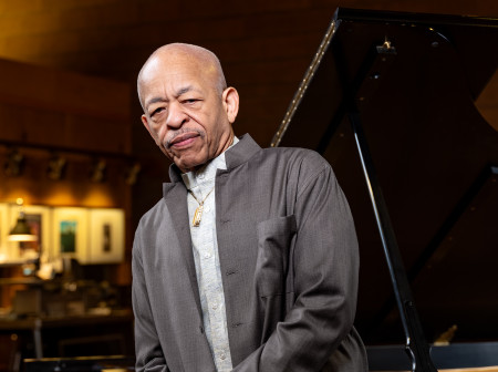 Steve Colson wears a gray suit and leans back against a Steinway grand piano.