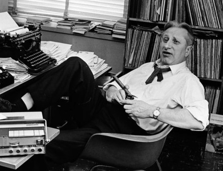 A grayscale image of Studs Terkel leaning back in an office chair surrounded by books and a typewriter.