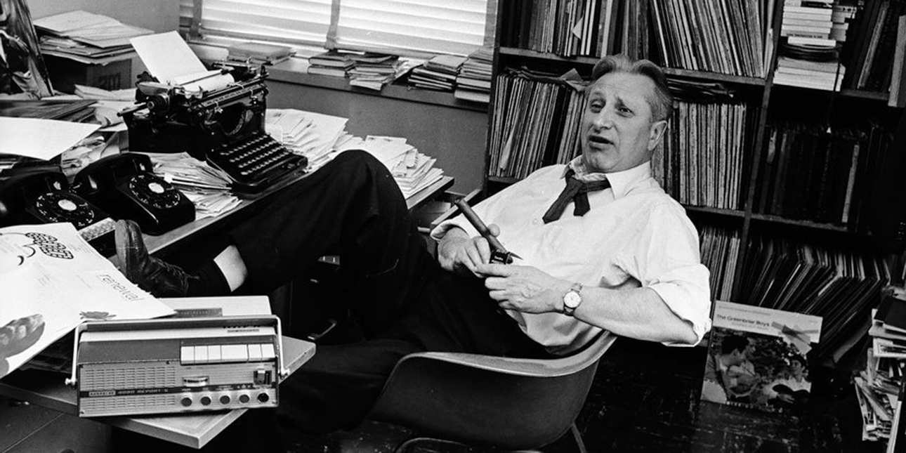 A grayscale image of Studs Terkel leaning back in an office chair surrounded by books and a typewriter.