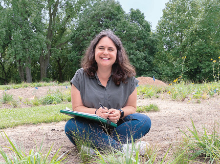 Becky Barak sits cross-legged in the dirt with a green clipboard on her lap and a pen in hand. Native plants can be seen in the forefront of the image, with tall green trees in the background.