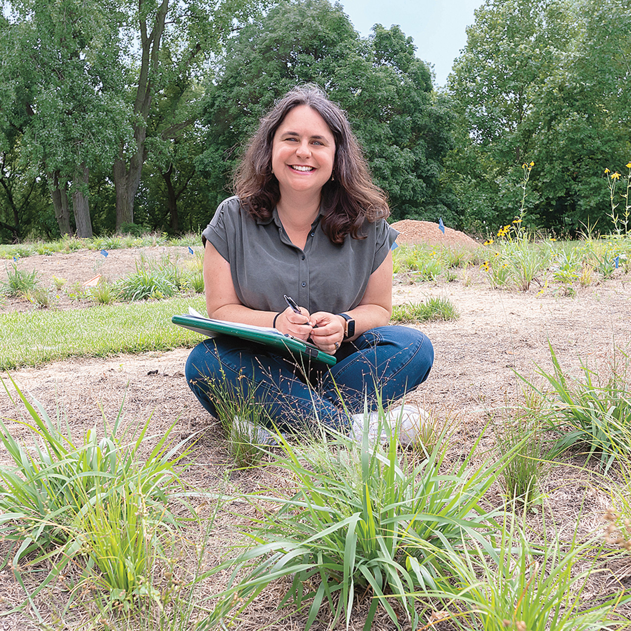 Becky Barak sits cross-legged in the dirt with a green clipboard on her lap and a pen in hand. Native plants can be seen in the forefront of the image, with tall green trees in the background.