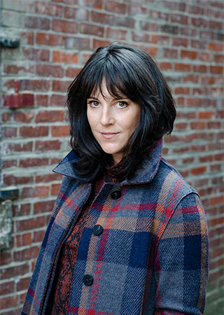 Amanda Dunlap poses for a headshot wearing a plaid peacoat in front of a brick wall.