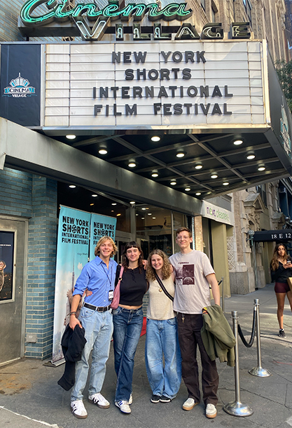 Four students stand next to each other at the Cinema Village in New York City. Above them, a marquee reads New York Shorts International Film Festival.