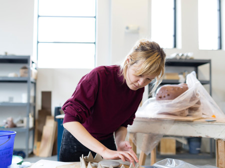 Lilli Carré wears a red sweater and leans over a workbench in her artist studio, working with clay.