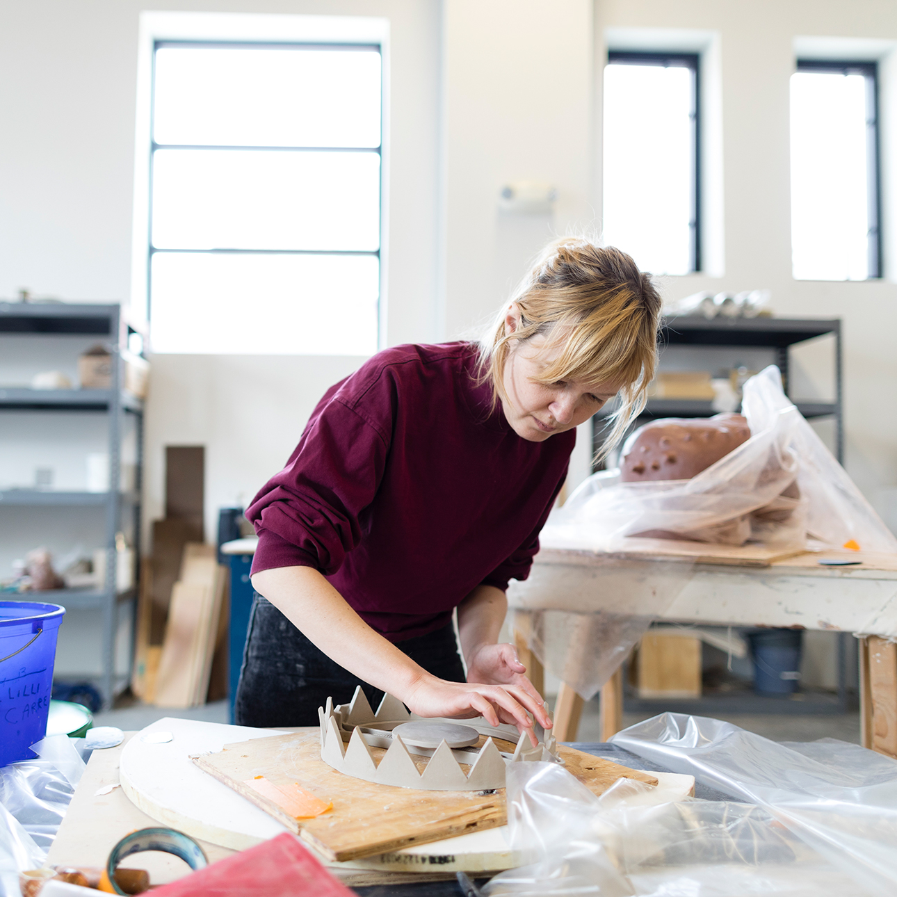 Lilli Carré wears a red sweater and leans over a workbench in her artist studio, working with clay.