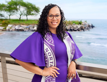 Lindsay Vahl Dean rests her arm on a railing overlooking Lake Michigan and smiles at the camera. She wears a purple dress and a Northwestern scarf.