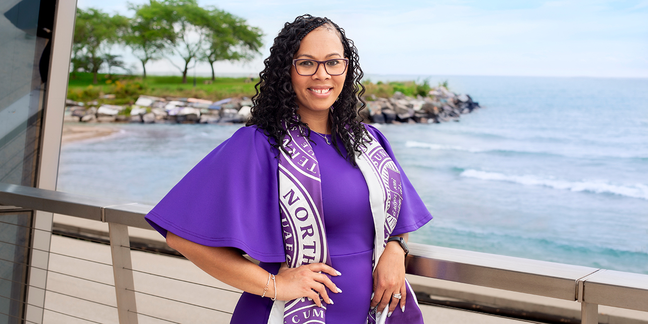 Lindsay Vahl Dean rests her arm on a railing overlooking Lake Michigan and smiles at the camera. She wears a purple dress and a Northwestern scarf.