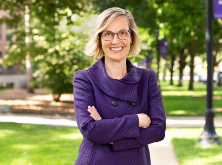 Kristin McDonnell is on campus, wearing a purple suit and crossing her arms while she smiles at the camera. Behind her are trees and a purple Northwestern banner hanging on a lamppost.