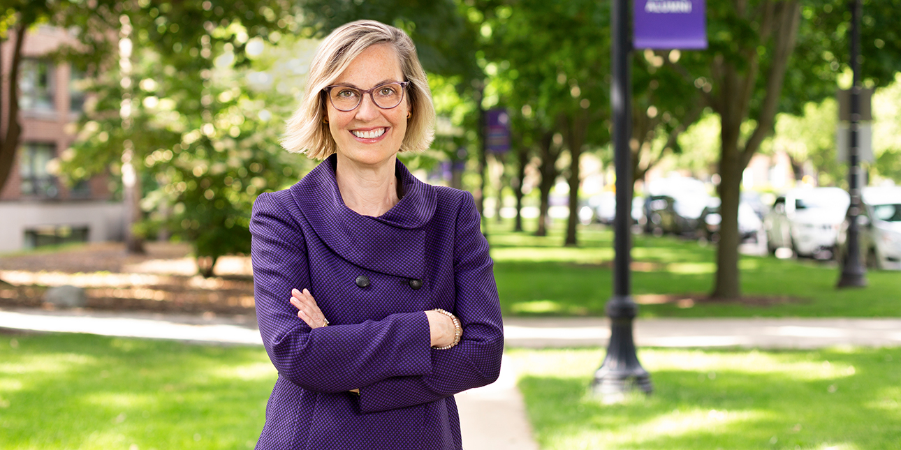 Kristin McDonnell is on campus, wearing a purple suit and crossing her arms while she smiles at the camera. Behind her are trees and a purple Northwestern banner hanging on a lamppost.