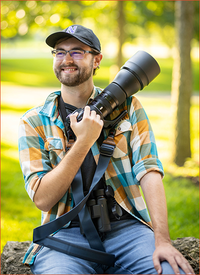 Collin Porter sits on a boulder on campus on a sunny day. He is wearing a blue-white-and-orange plaid collared shirt, blue jeans and a black baseball cap with the Northwestern N on it. He wears glasses and smiles while looking off into the distance. He is holding a professional camera across his chest and also has binoculars in his lap.