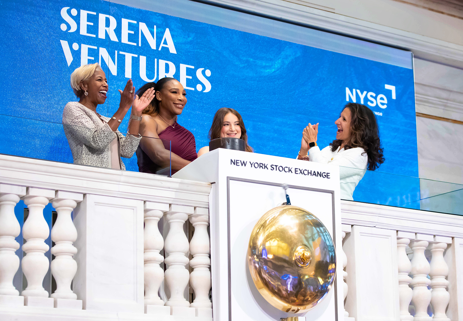 Sharon Bowen grins and claps her hands alongside Serena Williams and Alison Stillman, who are smiling, and NYSE Group president Lynn Martin, who is also smiling and clapping. The four of them stand behind a white dais, which is labeled with the words “New York Stock Exchange” and also boasts a large ringing bell. Behind them is a blue background displaying the words Serena Ventures and NYSE.