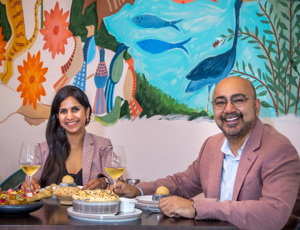 Ajit and Sukhu Kalra smile at the camera while sitting at a table at their restaurant, Indus Progressive Indian. The wall behind them is painted with birds, fish, flowers and other wildlife.