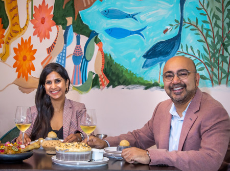 Ajit and Sukhu Kalra smile at the camera while sitting at a table at their restaurant, Indus Progressive Indian. The wall behind them is painted with birds, fish, flowers and other wildlife.