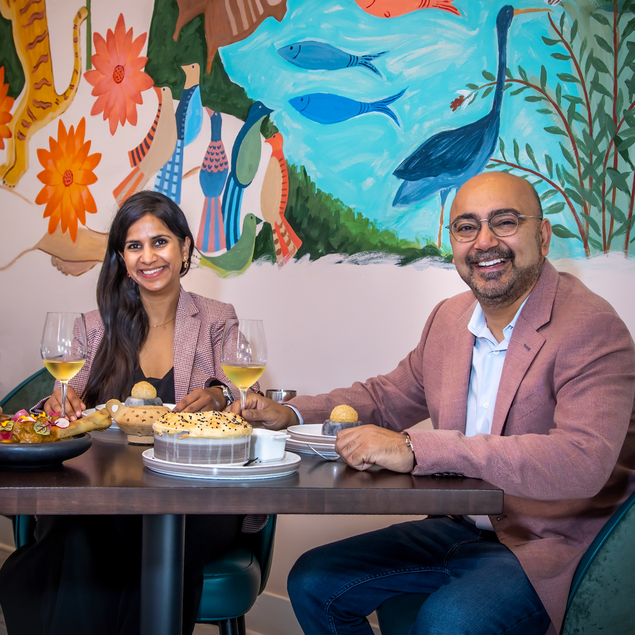 Ajit and Sukhu Kalra smile at the camera while sitting at a table at their restaurant, Indus Progressive Indian. The wall behind them is painted with birds, fish, flowers and other wildlife.